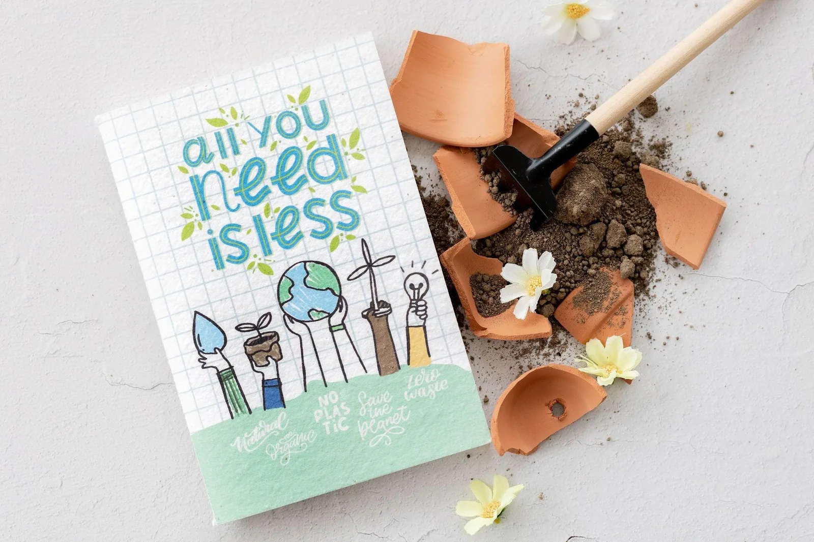 BĪJ notebook titled All You Need Is Less beside broken terracotta and soil